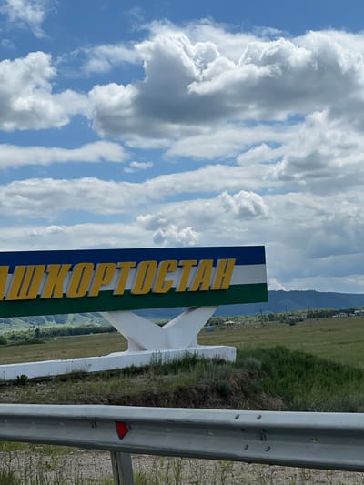 The image shows a sign with yellow letters spelling "БАШКОРТОСТАН" (Bashkortostan) against a backdrop of a blue sky with white clouds. The sign is mounted on a white structure. Below the sign, there is a green field with some trees and distant hills. The foreground shows a metal guardrail with a red reflector. The scene appears to be a roadside entrance to a region or city, possibly in or near Александровка, Russia. There are no people visible in the image.