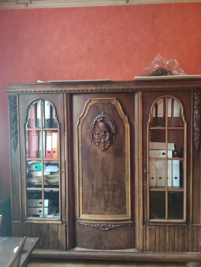An antique wooden cabinet stands against a coral-colored wall in what appears to be an office or study. The cabinet is ornate, with carved details on its doors and sides. It has three sections, each with glass-fronted doors revealing shelves filled with binders and papers. The left section contains colorful binders. The central section features a carved crest. The right section holds white binders and more papers. The room is dimly lit, suggesting an indoor setting. The image is taken from a medium distance, capturing the entire cabinet. No people are visible in the image. The scene does not provide any specific indicators of its location within Dresden, Germany.