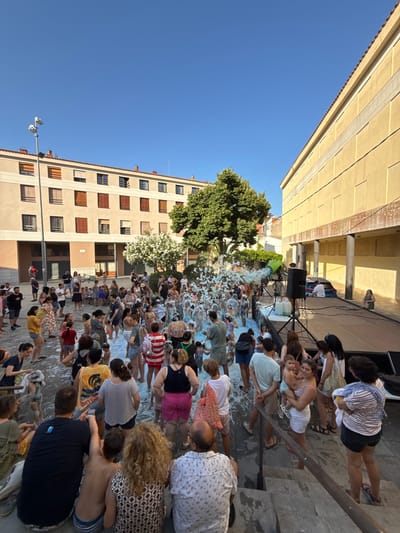 In Tossa de Mar, Spain, a foam party is in full swing in a town square. A large crowd of people, both adults and children, are gathered in the square, many of them immersed in a mound of white foam. A foam machine is visible, actively spraying foam into the area. In the background, there are buildings with distinctive architecture, typical of Spanish towns, and a clear blue sky overhead. Some individuals are standing on a raised stage, and a car is parked behind the stage. The scene suggests a lively community event on a sunny day.