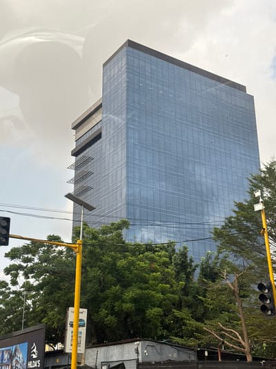 This image depicts a multi-story building with a blue-tinted glass facade in Falomo, Nigeria, under an overcast sky. The building exhibits a dark flat roofline and features several horizontal louvers on its upper left side. The glass reflects the cloudy sky and portions of its surroundings.

In the foreground, dense green trees partially obscure the building's base. Two prominent yellow poles are visible: the pole on the left supports a black three-lens traffic light fixture, while the pole on the right supports another similar traffic light fixture and a white, rectangular solar-powered street light or surveillance camera unit mounted at its apex. Overhead power lines traverse the mid-ground.

At the lower left, a partial black billboard displays the word "HILDA'S," and a smaller white sign with "LAGOS" is visible behind the left yellow pole. Below the tree line and behind the signage, low-rise dark structures and fencing are present. The sky is largely grey and cloudy, with a small dark object, possibly a bird, faintly visible in the upper right portion of the sky. No individuals or specific actions are discernible within the frame.
