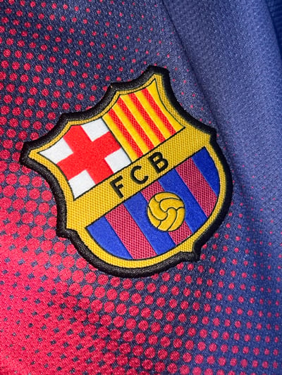 The image displays a close-up of a FC Barcelona crest embroidered on a blue and red dotted fabric, likely a sports jersey. The crest features the traditional red and white stripes, yellow and red stripes, blue and red stripes, and a yellow football. The fabric exhibits a textured pattern of red dots on a blue background in some areas, transitioning to a solid blue fabric in others. The crest is detailed and appears to be in good condition. There are no people visible in the image, and the environment is solely defined by the fabric and the crest. The location of Villa de Cura, Venezuela, is not depicted or suggested by the visual information present in the image.