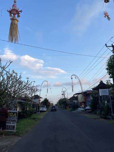 A street scene in Tabanan, Indonesia, with a road running through a residential area. Tall, decorative structures, likely for a festival or religious observance, hang from wires overhead and are erected on either side of the road. These structures feature intricate designs and tassels. The sky is blue with scattered white clouds. On the left side of the road, a sign is visible with text that includes "IMPORT" and "ARUM". A person is partially visible on the far left, interacting with the sign. Houses with traditional Balinese architecture line the street, some with decorative elements on their roofs. A white car and a dark-colored car are parked or driving on the road. Trees and greenery are present along the roadside. Overhead power lines are strung across the scene.