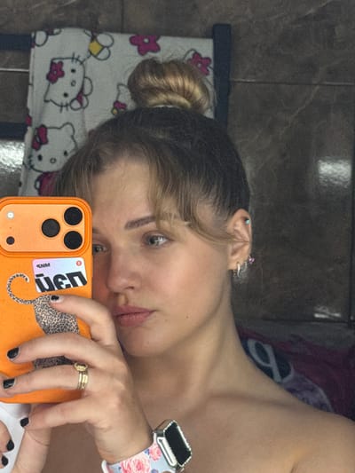 A close-up, rotated selfie captures a young woman's reflection in what appears to be a mirror or glass surface, taken indoors in Большая Шильна, Russia. The woman, with light brown hair styled in a bun and bangs, looks directly towards the camera. Her right ear is visible with multiple piercings adorned with small, colorful studs.
She is wearing a sleeveless top or bare-shouldered attire, revealing her collarbone and shoulder. On her right wrist, she wears a smartwatch with a decorative white band featuring pink and blue floral patterns. A gold ring with a central stone or design is visible on her right ring finger. Her fingernails are painted with dark polish.
The woman holds an orange smartphone with a triple camera lens array in her right hand. The phone case features a sticker with a leopard print design and partially visible reversed text: "NGJu" and "MNS", along with a red "R" logo. A white blanket with Hello Kitty characters and pink flowers is visible beneath her head, suggesting a casual, home environment. The background consists of a dark, mottled, possibly tiled wall. The overall scene depicts a casual selfie being taken.