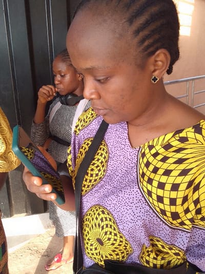 The image depicts three individuals in an outdoor or brightly lit indoor setting in Nkwelle Ezunaka, Nigeria. In the foreground, a woman with cornrow braids and dark skin is shown in profile, facing right, looking down intently at a dark green cased smartphone held horizontally in her right hand. She wears a v-neck top with a light purple background featuring small dark dots and large yellow and black geometric patterns. A black strap crosses her chest, likely from a bag. Her left ear displays a black clover-shaped earring with a gold border. Behind her, a second woman, slightly out of focus, has dark hair pulled back and wears a grey and white patterned top. Black over-ear headphones rest around her neck. Her right hand is raised towards her mouth, and her left foot, wearing a red sandal with a pink decorative element, is visible. A portion of a third person in yellow patterned clothing is partially visible on the far left. The background features dark vertical paneling behind the second woman and a light-colored railing to her right. The ground is a light-colored, possibly concrete or dusty surface. Strong lighting illuminates the scene.