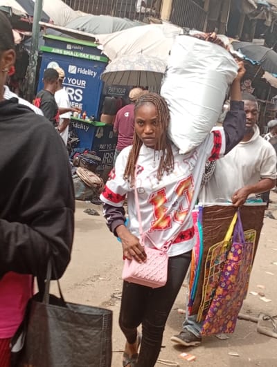 In a bustling outdoor market, a woman carries a large white bag on her shoulder, looking towards the camera. She wears a white shirt with red and black accents, black pants, and a small pink purse. To her right, a man carries a stack of colorful bags. Behind them, a Fidelity AIRS SMALL-TAX HUB is visible with promotional text. Umbrellas and crowds fill the background, indicating a busy commercial area.
