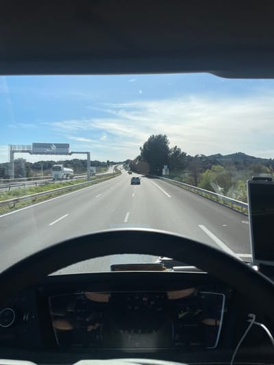 The image depicts a forward-facing view from inside a vehicle traveling on a multi-lane highway in Tavel, France, under a clear blue sky with scattered white clouds. The lower third of the frame is occupied by a dark-colored steering wheel and a portion of the vehicle's digital instrument cluster, which displays a speed of approximately 70-80 km/h. A dark object, possibly a wallet, rests on the dashboard in the center. To the right of the steering wheel, a dark electronic device, likely a smartphone, is mounted, with a white charging cable extending downward.
The highway is an asphalt road with white dashed lane markings, showing at least two lanes of travel in the depicted direction. Ahead in the right lane, a light-colored car is visible, followed by a darker vehicle further in the distance. In the left lane, a white tanker truck is also traveling forward. An overhead gantry structure spans the highway on the left, supporting several unreadable rectangular signs. Metal guardrails line both sides of the road. The environment surrounding the highway features green trees and bushes, with low hills visible on the horizon to the right. The scene is illuminated by bright daylight.