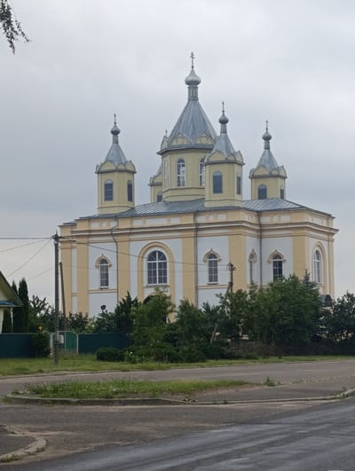 The image shows a large, ornate yellow and white church with multiple domes and a cross on top, located in Дзярэчынскі сельскі Савет, Belarus. The church has a prominent central dome and several smaller domes around it, all topped with silver roofs and crosses. The building's facade features a combination of yellow and white paint, with arched windows and decorative trim.
In front of the church, there is a road and a grassy area. A utility pole with wires runs alongside the road, partially obscuring the view of the church's lower section. Behind the church, a dense line of green trees is visible. The sky is overcast, suggesting a cloudy day. A small portion of a house with a light-colored roof and green siding is visible on the far left of the image. The overall scene depicts a place of worship within a semi-rural or suburban setting.