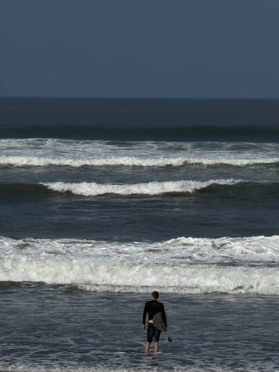 A male individual stands waist-deep in the ocean water, facing away from the viewer towards the horizon in Belalang, Indonesia. He possesses short, reddish-brown hair and wears a black long-sleeved wetsuit top and dark shorts. A light-colored surfboard with a black leash is held under his left arm. The ocean water is dark blue-grey with several white-capped waves breaking and rolling towards the shore. The sky above is a solid, dark grey-blue.