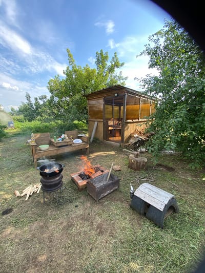 The image depicts an outdoor scene in the village of Kemul, Russia, on a clear day with scattered clouds. The foreground features a fire pit with active flames, a large black cauldron on a stand cooking food, and a wooden bench with various items on it, including bowls and bottles. To the right of the fire pit is a makeshift structure constructed from tires and wood. In the background, a wooden and translucent-sided structure, possibly a gazebo or greenhouse, is visible. Lush green trees and vegetation surround the area, and in the far left, a greenhouse structure can be seen. The ground is covered in grass and dirt.