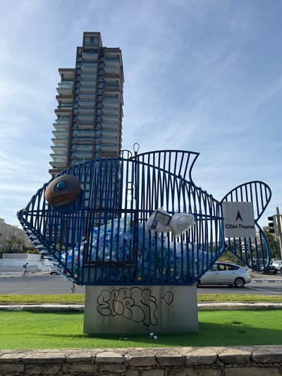 A daytime, outdoor photograph depicts a blue fish-shaped sculpture bin for plastic bottles, located in Germasogeia, Cyprus. The bin is partially filled with clear plastic water bottles and a few pieces of white litter. Attached to the top is a sign that reads "OSM Thome." The bin is mounted on a grey metal post bearing graffiti. A single, brown, circular eye is affixed to the side.
To the left of the bin is a tall, modern residential building with staggered balconies. The sky is light blue with wispy clouds. To the right of the bin is a paved road with a parked white van, a narrow strip of green grass, and a stone wall. Part of another building and a traffic light are visible in the background. In the distance, two people can be seen on the sidewalk. The photograph appears to be taken from a slightly low angle.