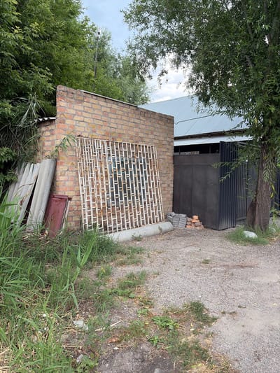 In the city of Kant, Kyrgyzstan, a close-up shot captures a scene near a brick building with a corrugated metal roof. In the foreground, tall green grass and some dry vegetation grow on a gravelly ground. To the left, several flat, gray construction panels lean against the brick building. Adjacent to them is a dark red rectangular object.
Dominating the center of the frame is a large, white, decorative metal grate, showing signs of rust, leaning against the brick wall. Behind the grate, a window with blue panes is visible in the brick structure.
To the right of the brick building, a tall, dark, ribbed metal gate stands closed. In front of this gate, a small stack of bricks is piled on the gravel. A mature tree with a thick trunk and green foliage grows to the right of the gate. The background reveals the sloped, light-colored metal roof of another building extending beyond the brick structure. The sky above is partly cloudy, with a light pole topped with a white object visible amongst the trees in the upper left portion of the image.