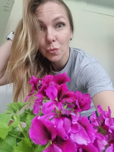The image depicts a fair-skinned woman with long blonde hair posing with a bouquet of bright pink flowers. She is wearing a gray t-shirt and a watch. The background appears to be a light-colored wall. There is no indication of the location being Kemul, Russia, or any urban environment.