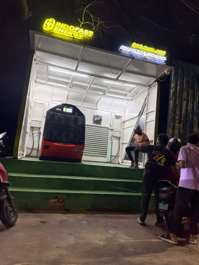 The image depicts an outdoor scene at night, likely in Chennai, India, featuring a modern electric scooter battery swapping station. The central focus is a white, open-air structure with a translucent roof, illuminated by bright white lights. Inside, a sleek, orange and black battery swapping unit is prominently displayed. It has a digital screen on its front and appears to be part of an electric mobility service.
Several people are present, mostly obscured by the low lighting and the angle of the shot. One individual in a black t-shirt with a yellow logo is leaning towards the swapping unit, while others stand nearby, some wearing helmets, suggesting they are scooter users. One person in the foreground wears a light purple shirt and black pants. The overall atmosphere is one of urban activity and technological advancement.
Visible text on illuminated signs identifies the business as "INDOFAST ENERGY," a "SUN Mobility Joint Venture," and indicates the service as a "SWAP POINT." The Tamil script alongside "SWAP POINT" translates to "Swap Point." Another sign reads "MMOBILITY" and "Hoppiness," with a logo that includes "%" and "Apollo." A GSTIN number is also visible. The setting appears to be a street or parking area, with a concrete ground and some greenery visible to the right. The presence of illuminated signs and artificial lighting confirms it is nighttime.
