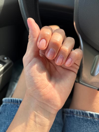 A person's hand, with natural-colored, short, rounded nails, is held up in front of the steering wheel of a car. Sunlight casts shadows across the skin. The person is wearing blue denim. The scene is set indoors, likely within a vehicle, and no specific location in Ust-Ilimsk, Russia can be identified.