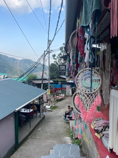 The image depicts a street scene in Bhagsu, India, with a narrow cobblestone path leading uphill. On the right side of the path, a market stall displays various handmade items, including dreamcatchers with white and pink tassels, macrame wall hangings, and colorful textiles. A bright red ledge separates the stall from the path, with folded fabrics resting on it. A partially visible white curtain hangs on the far right. In the background, there is a sign for "BABA CAFE BHAGSU - HIMPI". Several people are visible; two men are sitting on the curb in front of the stall, and two men are seated at a table outside a cafe on the left. The cafe has a green tin roof and a pink exterior wall. Beyond the cafe, the cityscape of Bhagsu unfolds, with buildings and rolling green hills under a partly cloudy blue sky. Numerous electrical wires crisscross the scene, strung between poles.