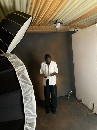 A Black man stands in a photo studio in Katon Rikkos, Nigeria, looking down at his hands. He is wearing a white short-sleeved button-up shirt and black pants with cargo pockets. He has dark hair and is wearing glasses. The studio features a dark backdrop, a large softbox to the left, a large white styrofoam board to the right, and draped yellow and cream fabric on the ceiling. A small light fixture is mounted on the ceiling. The floor is concrete.