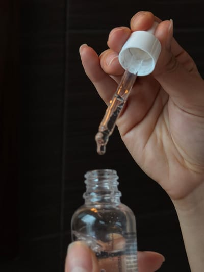 A person's hand holds a clear, small bottle with ribbed neck. The bottle is partially filled with liquid. The other hand holds a dropper with a white cap. The dropper contains a clear liquid and a single drop hangs from the end, positioned near the bottle's opening. The background is a dark, textured surface.