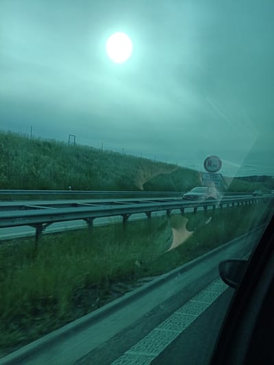 The image presents a view from the passenger window of a vehicle traveling on a highway near Třemošnice, Czechia. The entire scene is characterized by a strong teal or greenish color cast, which could be due to window tinting or atmospheric conditions.
In the upper left, a bright, intense white orb, likely the sun, is prominent against a hazy, greenish sky. Below this, a grassy embankment slopes upwards, topped by a metal fence. Reflections on the car window are significant, superimposing blurred shapes from the car's interior, such as parts of the dashboard or a hand, onto the landscape. These reflections are particularly noticeable over the grassy areas and the road surface, creating an almost double-exposure effect.
The highway occupies the mid-ground, featuring multiple lanes separated by metal guardrails. Another dark-colored vehicle is visible ahead on the highway. On the far right, a black car side mirror and a portion of the car's interior door frame are visible in the foreground.
A roadside sign is clearly visible on the right side of the highway. It displays a circular regulatory sign with a red border and a diagonal line, which typically indicates a prohibition, possibly for trucks. Below this, the text "6-22h" suggests the hours of the restriction, followed by "200 m," likely indicating the distance to the restriction or a specific point. The setting suggests daytime travel, likely under overcast or hazy conditions given the diffused appearance of the sun and the overall greenish ambiance.