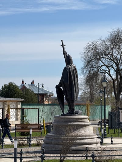 The image captures a bright, clear day in an outdoor public space, likely a park or square, in Ново-Никольское, Russia. The central subject is a dark-colored statue of a historical figure, possibly a warrior, standing prominently on a light-colored, fluted stone pedestal. The figure wears a helmet and a flowing cloak, holding a sword pointed upwards in one hand and seemingly a shield or similar object in the other.
In the upper part of the frame, a large, deciduous tree with bare branches spreads across the blue sky, suggesting either early spring or late autumn. The background reveals buildings with traditional architectural styles, featuring peaked, possibly metallic, roofs, some with chimneys and decorative finials. A green corrugated fence or wall is also visible behind the structures.
The foreground and mid-ground are furnished with typical public amenities, including paved pathways, green grass, and scattered small bushes. Several ornate black street lamps, each with multiple lanterns, are strategically placed throughout the area, alongside wooden park benches. In the lower right corner, a person dressed in a dark jacket and blue jeans is visible, appearing to be resting on or leaning against a barrier. The scene conveys a sense of calm and order under a bright sky with a few wispy clouds. No visible text is discernible in the image.