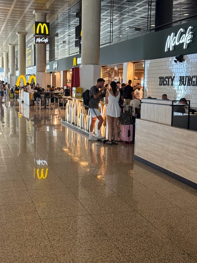 This image depicts a scene inside a McDonald's restaurant, likely located in an airport terminal at El Prat de Llobregat, Spain, given the presence of numerous travelers and the polished, spacious interior indicative of such a location.
In the foreground, a man wearing a grey t-shirt, white shorts, and white socks with grey slides is standing, facing to the right. He has a backpack over his right shoulder and appears to be holding something in his right hand, possibly food or a drink. To his left, a woman in a white dress with thin straps and black sandals is standing, facing towards the man. She has braided hair and is looking down, holding a white object in her left hand. Between them, a pink suitcase with black wheels is placed on the floor.
Behind them, a counter area is visible with several people queuing or sitting at tables. A sign with the "McCafé" logo is prominent above the counter. Further back, more seating areas and patrons are visible, along with other McDonald's branding. The floor is highly reflective, showing the illuminated McDonald's "M" logo from the ceiling as a bright yellow reflection.
The environment appears to be an indoor public space, likely a commercial area within a transportation hub. The lighting is bright, and the architecture features large pillars and a high ceiling with visible structural elements. The overall atmosphere is one of a busy commercial establishment catering to travelers.