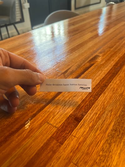 A hand holds a fortune cookie slip with the message "Malas decisiones hacen buenas historias" (bad decisions make good stories). The slip also has "RWR Café Club" printed on it. The image appears to be taken indoors at a cafe in Avellaneda, Argentina, indicated by the "RWR Café Club" being located in Avellaneda. The background is a wood-grained table. A cup and some chairs are visible in the blurred background.