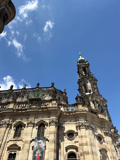 The image shows the Dresden Frauenkirche in Germany from a low angle, showcasing its ornate Baroque architecture against a blue sky with scattered clouds. The sandstone exterior of the church is detailed with numerous sculptures and architectural elements. A banner hangs on the side of the building. The top of the dome is visible, capped with a cross and a lantern. Scaffolding is present, suggesting ongoing restoration work.