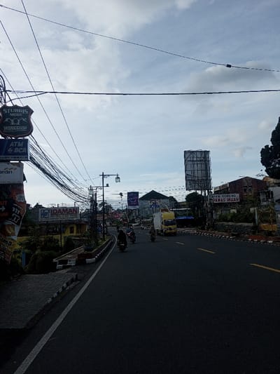 The image depicts an urban street scene on a cloudy day.  Multiple overhead wires crisscross the sky above the road.  Signs for "Sturgis Moge Workshop", "ATM BCA", and "Idaman Hotel Cottage" are visible, along with a large billboard and the signage "Hayam Goreng Panas Indah".  Several motorcycles and a yellow truck travel down the road, suggesting moderate traffic. A sidewalk borders the road on the left.