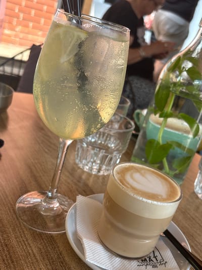 This media file captures a close-up of a table at an outdoor cafe, likely in Kragujevac, Serbia, given the context. The scene features two drinks and a bottle of water with plants.
In the foreground, a stemmed glass filled with a bubbly, pale yellow beverage, possibly a spritzer or lemonade, is prominently displayed. The glass is covered in condensation, suggesting it's a warm day. Two black straws are visible in the drink. Behind this glass, a transparent glass with a textured surface, possibly a tumbler, sits empty. Further back, a clear glass bottle contains water with green plant stems and leaves submerged in it. To the right, a clear glass mug holds a frothy coffee drink, likely a latte or cappuccino, with latte art visible on the surface. This mug rests on a small white saucer with a white napkin. The napkin has a printed logo that appears to say "MAT" in stylized lettering, with "espreso bar 2017" below it, along with a small graphic element.
The background is blurred, showing glimpses of people and what appears to be an outdoor seating area with reddish-brown tile or brickwork. The lighting suggests it could be daytime, possibly late morning or afternoon, given the brightness and the condensation on the glass. The overall atmosphere seems relaxed and casual, typical of a cafe setting. The image focuses on the beverages and the immediate table setting, with hints of the surrounding environment. No specific emotions are directly conveyed, but the presence of drinks and the cafe setting imply a moment of leisure or social gathering.