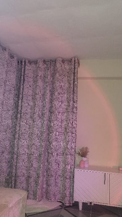 An indoor scene in Nanyuki, Kenya, is depicted, illuminated by a pervasive pink and purple light, originating from a source creating a distinct arc on the right wall. The left side of the frame is occupied by drawn, floor-length curtains featuring a grey and purple abstract geometric pattern, hung on a rod with grommet rings. To the right, a light-cream wall displays a horizontal molding. A white, two-door cabinet with chevron-patterned fronts and four light-colored legs stands against this wall. Upon the cabinet are a white vase containing dried foliage and two small pink cylindrical decorative items. A portion of a light-colored, upholstered furniture item is visible in the bottom-left foreground. The floor consists of dark geometric tiles, partially covered by a light-colored rug positioned beneath the curtains. No individuals are present.