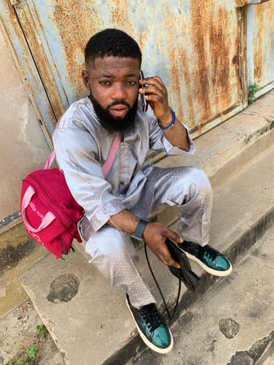 An adult male is seated on weathered concrete steps in Okene, Nigeria. He wears a light grey two-piece garment with a subtle dotted pattern, black and teal patent leather sneakers with white soles, a black smartwatch on his left wrist, and a blue beaded bracelet on his right wrist. A red "Hello Kitty" brand bag with a pink strap is slung over his left shoulder. He holds a black smartphone to his right ear with his right hand, indicating a phone conversation. His left hand rests on his left thigh, holding a black wallet or phone case with a strap. Behind him is a large, severely rusted metal surface, likely a gate or door, exhibiting extensive brown and orange oxidation. A small green plant grows from a crack in the concrete near the rusted surface on the right side of the frame.