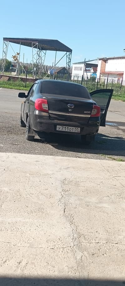A black Datsun sedan is parked on a gravel surface in front of a concrete curb. The car's driver-side rear door is open. The license plate of the car is visible and reads "Y 711 OT 55". In the background, there is a large metal structure with a dark roof, a fence, and several buildings, suggesting an urban or semi-urban environment. The sky is clear and blue. The scene appears to be in Okoneshnikovo, Russia, as indicated by the license plate's region code "55".