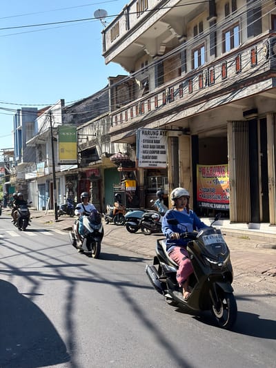 The image depicts a street scene in Buleleng, Indonesia, on a sunny day. Multiple motorcycles are navigating the asphalt road, which is marked with a pedestrian crossing. On the right side of the road, there is a building with multiple levels and shuttered storefronts. A prominent banner on one of these storefronts advertises "WARUNG AYE" and lists various food items, including "SYOBAR BABI," "NASI BABI GULING," "NASI CAMPUR BABI," and "BAKSO BABI," along with an address. Further down the street, another banner for "WARUNG AY..." is visible. To the left, a building with a blue facade features an "OPPO" and "Realme" sign, along with "JK PHONESHOP." Overhead, electrical wires crisscross the sky, and a satellite dish is mounted on the upper level of a building. People are riding on the motorcycles, most wearing helmets for safety. The lighting suggests it is daytime, with strong shadows cast by overhead wires and the buildings.