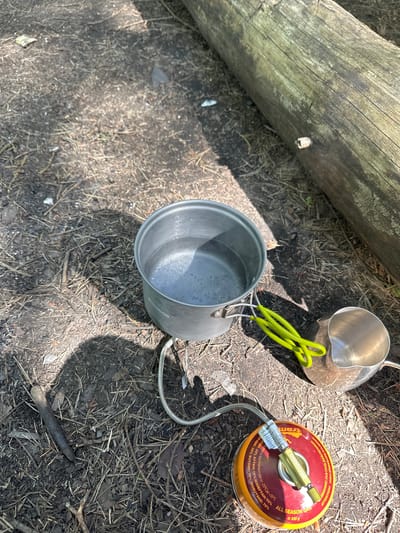 The image shows camping equipment laid out on the ground in what appears to be a forested area, likely in Юзуфоўскі сельскі Савет, Belarus. A log is visible in the upper left of the frame. Two metal camping pots and a fuel canister with a stove attachment are arranged on the forest floor, which is covered in dirt, pine needles, and small twigs. One of the pots has green handles. The fuel canister is red and yellow and has writing on its surface. The stove attachment is connected to the canister via a flexible metal hose. The scene suggests someone is preparing to cook or has recently cooked while camping.