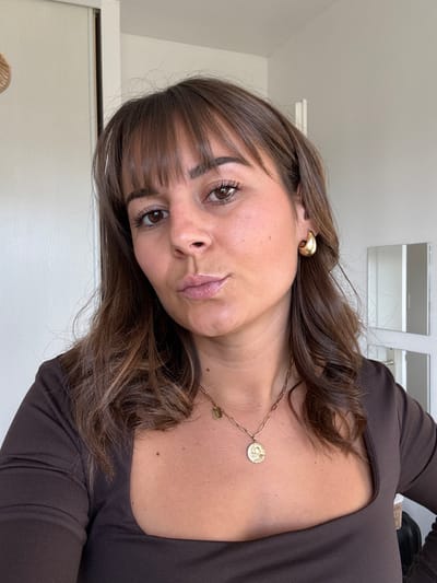 The image depicts an adult female posing for a close-up photograph indoors, located in Veauchette, France. The subject has long, wavy brown hair with bangs covering her forehead, brown eyes, and a tanned complexion. Her lips are slightly parted and puckered. She wears a dark brown long-sleeved top with a square neckline. A gold-colored hoop earring is visible on her right ear. Around her neck are two gold-colored necklaces: a paperclip chain with a prominent round medallion pendant featuring an embossed design, and a finer chain with a smaller oval pendant. The background consists of a light-colored wall with a vertical seam or door frame on the left. On the right, reflective panels with white framing are visible, suggesting a mirror or closet doors. A partially visible, light-colored woven circular object hangs on the upper left wall.