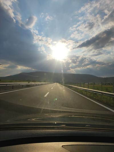 The image shows a view from inside a vehicle, looking out at a highway in Greece. The sun is bright and partially obscured by clouds, suggesting it is either sunrise or sunset. The sky is a mix of blue and grey clouds, with sunbeams breaking through. The highway stretches ahead, with a white line marking the lanes. There are guardrails on either side of the road, and beyond them, fields and a gently sloping hill. Power lines are visible on the hill in the distance. The overall mood is peaceful and serene, with the warm sunlight creating a sense of calm. The scene is likely taking place during the golden hour, adding to the visual appeal of the landscape. The location is identified as Farkadona, Greece. There is no visible text in the image.