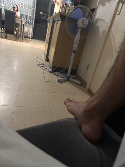 A dimly lit indoor scene shows a person's bare foot resting on what appears to be dark grey fabric, possibly pants, in the foreground. The foot is positioned towards the bottom right of the frame. The background reveals a tiled floor, a white electric fan with blue blades, and a beige cabinet with multiple drawers. To the left, a blurred figure of a woman is visible sitting at a table and looking at her phone. The overall environment suggests a domestic setting, likely a dining area or living space, in Waregem, Belgium.