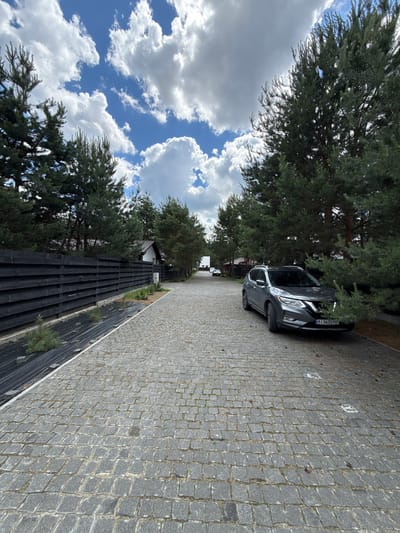 The image depicts a scene in Khmilna, Ukraine, on a bright, cloudy day. The perspective is from ground level, looking down a paved driveway lined with trees and a dark wooden fence. In the foreground, a grey SUV is parked with its hood up, suggesting some sort of mechanical issue or inspection. The driveway is paved with cobblestones, some of which are marked with white squares, possibly indicating parking spots.

Behind the fence, a white house with a brown roof is visible, nestled amongst tall evergreen trees. The sky is a vibrant blue, punctuated by fluffy white clouds, indicative of fair weather. The overall atmosphere is one of a quiet, suburban or rural setting. The license plate of the car is partially visible, displaying "AI 5635 PP". The orientation of the image is such that the scene appears to be viewed from a side angle, with the driveway stretching into the distance.