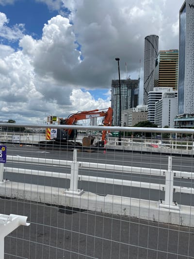 The image captures an urban construction scene in South Brisbane, Australia, viewed from an elevated perspective, likely a bridge or elevated walkway. In the foreground, a temporary metal mesh fence separates the viewer from a multi-lane road or bridge undergoing construction or maintenance. White barriers and supports line the edge of the road, which appears to be dark asphalt with white lane markings.

Two orange excavators are visible within the construction zone to the left of the road. The larger excavator is positioned with its arm extended downwards, its digging bucket resting near an excavated area, suggesting ongoing or recent work. The smaller excavator is partially visible behind it. Text such as "HITACHI" and the "D&R PLANT HIRE" logo are legible on the machinery, along with a yellow tag displaying numbers like "142". A tall, dark pole with a spherical top stands upright near the excavators.

Beyond the immediate construction area, a dense cityscape fills the midground and background, featuring numerous modern high-rise buildings under varying stages of completion. Several tall cranes indicate active building construction. One prominent, cylindrical skyscraper exhibits a distinctive helical or striped pattern. Other buildings display legible text, including "VOCO" and "QUEENSLAND". The sky above is partly cloudy, with large, fluffy cumulus clouds interspersed with patches of bright blue, indicating daytime conditions. A blue sign attached to the fence in the lower left corner prominently displays "D&R PLANT HIRE" and its website, "DRPLANTHIRE.COM.AU". The overall scene depicts a dynamic urban environment characterized by significant ongoing development.
