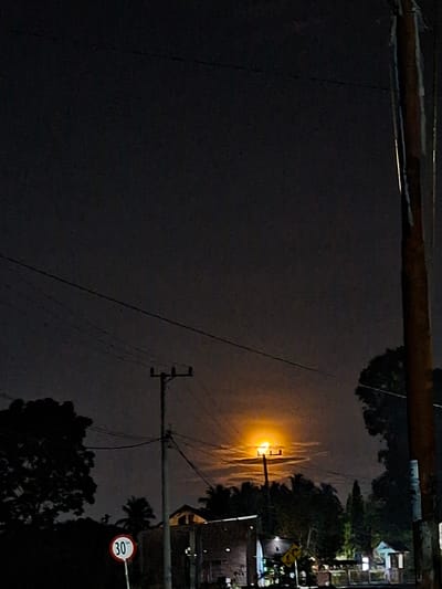 The image shows a night scene in Kutaglumpang, Indonesia. A street lamp illuminates the area, casting a bright orange glow. Power lines crisscross the sky. A speed limit sign reading "30" is visible on the right. There are utility poles, trees, and silhouettes of buildings. The sky is dark and overcast. The perspective is tilted, with the foreground dominated by darkness.