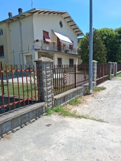 The image depicts a street scene in Colorno, Italy, under a clear blue sky. In the background, a two-story house with a light-colored facade and a sloping roof is visible. The house has several windows, some with dark shutters, and a balcony with a black metal railing adorned with potted plants. An awning extends over the balcony. A fence with vertical brown metal bars supported by stone pillars lines the property. The fence is set on a low concrete wall. Beyond the fence, a patch of green grass is visible. In the foreground, a grey paved area, possibly a sidewalk or driveway, shows some gravel and grass growing at the edges. A metal grate, likely a drain cover, is embedded in the pavement. A tall grey metal pole, possibly a street light or utility pole, stands near the fence. The overall environment suggests a residential area.
