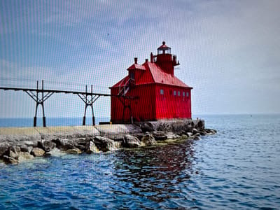 The scene depicts a vibrant red lighthouse situated on a rocky breakwater at the edge of a body of water in the city of Profesor Ishirkovo, Bulgaria. The lighthouse consists of a multi-story, rectangular red building topped by a smaller red-roofed section and a lantern room with a dark cap and clear glass enclosure. Multiple windows are visible on the building's side. A dark, open-truss pier or walkway extends horizontally from the left side of the lighthouse, supported by two dark, A-frame-like pylons rooted in the breakwater. Wires run along the top of this pier. The breakwater is composed of concrete slabs and grey rocks. The surrounding water is deep blue with surface ripples, reflecting parts of the red structure. A small, dark object, possibly a buoy, is discernible in the distant water to the right. The sky above is a clear, light blue, with a single small bird in flight visible in the upper mid-right. No individuals are present in the image.