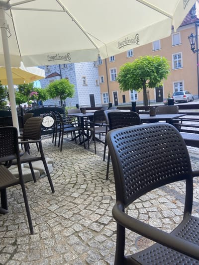 The image shows an outdoor seating area of a cafe or restaurant in Bolesławiec, Poland. The scene is captured from a low angle, looking up and across a cobblestone pavement. Several dark brown plastic chairs with a perforated design are arranged around dark tables. The chairs are lined up, suggesting the establishment is either closed or about to open, as there are no patrons visible.

In the background, there are buildings with yellow and light blue facades. One building has a distinctive geometric pattern on its facade. Trees with lush green foliage are planted between the buildings and the seating area, adding a touch of nature to the urban setting. A large white umbrella, partially visible in the foreground, provides shade over the seating area. The umbrella has a logo printed on it, which appears to be text in a decorative font, possibly advertising the establishment. Another smaller, yellow umbrella is also visible.

A sign with text is visible on one of the tables, featuring a circular emblem with the words "THE TAVERN" and other decorative elements. The time of day appears to be daytime, with bright natural light illuminating the scene, suggesting pleasant weather. The cobblestone street and the architecture of the buildings hint at a European location. There are no people visible in the image, and the overall atmosphere is calm and quiet. The focus is on the outdoor dining setup and the surrounding environment.