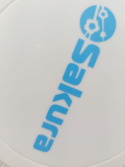 A white round object, possibly a lid or a plate, has the word "sakura" printed on it in blue. The letters are arranged diagonally from the bottom left to the top right. Above the letter "s" is a blue circle with stylized white keys inside it. The background is a plain white surface.