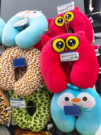 The image displays a retail store's vibrant selection of U-shaped neck pillows, likely taken indoors in Sindanglaya, Indonesia. The pillows are arranged on hooks against a light gray wall or display unit with a black grid pattern. There are multiple pillows with different designs: several bright red and light blue owl-themed pillows featuring large, cartoonish eyes; a pillow with a brown and white giraffe-like print; and another with a green spotted or camouflage pattern. A small portion of a leopard print pillow is also visible at the bottom left.
Each visible pillow has a price tag attached, uniformly displaying "Rp 42.000," which is 42,000 Indonesian Rupiah. The text "DB BANTAL LEHER U KU" (likely referring to "DB U-Shaped Neck Pillow") is prominent on the tags, along with barcodes and other smaller, less legible text like "TAM MILWA". The brand "J&L" is visible on some of the red owl pillows. The scene depicts a typical retail display, inviting customers to browse and purchase comfort accessories. No people are visible, but the implied activity is shopping.