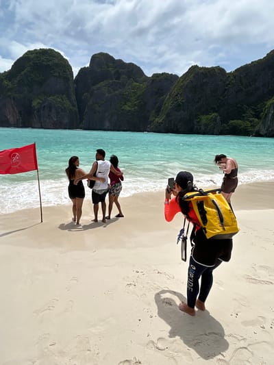 In Ao Nang, Thailand, a woman wearing a bright orange long-sleeved shirt and black pants, with a yellow waterproof bag slung over her shoulders, is photographing a group of three people standing near the water on a sandy beach. The group of three consists of a man and two women. The man is wearing a white tank top and dark shorts, with his arms around the shoulders of one woman who is wearing a black swimsuit. The second woman, wearing a maroon top and patterned shorts, stands beside them. A red flag with a symbol indicating no swimming is planted in the sand to their left. In the background, turquoise water meets a white sandy beach, backed by lush green, rocky cliffs under a cloudy sky. Another woman in a dark bikini top and shorts is seen further down the beach, facing away from the camera. The sand is marked with footprints and shadows.