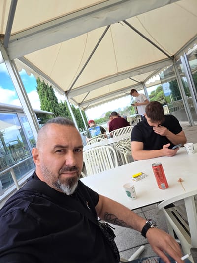 The image captures a selfie-style shot of a man in the foreground, taken from a slightly high, tilted angle. The man, with a shaved head and a graying beard, wears a black t-shirt and has tattoos on his left arm, one of which appears to spell "NILO." He is wearing a black wristwatch with a metallic face and is holding a lit cigarette in his left hand, which is resting on a white table.

On the white table in front of him are several items: a red aluminum can (likely a soda or energy drink), a white disposable paper cup with a green leaf logo, an open yellow and white pack of Marlboro Gold cigarettes, a yellow lighter, and a used wooden toothpick.

The scene is set under a large, light-colored tent or marquee structure with visible metal framework, suggesting an outdoor dining area or casual gathering space in Sant Cugat del Vallès, Spain. In the background, several other individuals are visible. One person, also wearing a black t-shirt, is seated at an adjacent white table, looking down at a phone. Further back, another person in a burgundy shirt is seated, and a person in a white t-shirt is standing, also seemingly occupied with a phone. A person wearing a red hat is visible in the middle background.

Beyond the tent, a clear blue sky with some white clouds and lush green trees are visible, reflecting in a glass building or partition. The overall atmosphere is casual and relaxed, likely during daylight hours, given the bright illumination. The primary activity appears to be people socialising, taking a break, and using their mobile phones.