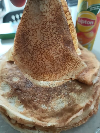 A close-up, slightly overhead shot focuses on a stack of crepes, with one crepe being held up by a finger in the foreground. The crepes are golden brown with a slightly lacy texture. In the background, out of focus, is a can of Lipton iced tea. The scene appears to be indoors, with a white tabletop and a blurred background suggesting a kitchen or dining area. There is no identifiable information in the image to suggest a specific location in Perpignan, France.