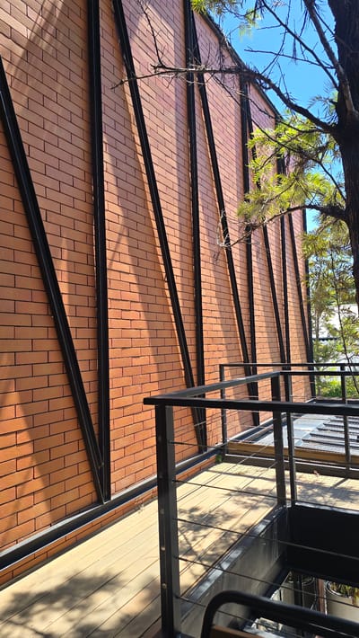 A low-angle, eye-level shot captures a modern building facade and an elevated wooden walkway. The facade is constructed from reddish-brown bricks and features a series of tall, dark metal support beams that extend vertically from the base to the top of the building. The beams create a striking geometric pattern against the brickwork, with shadows from branches and leaves casting dappled patterns across the wall. A tree with green foliage is partially visible in the upper right corner, its branches reaching towards the blue sky.
In the foreground, a wooden walkway with a light-colored plank surface runs parallel to the building. A black metal railing with thin horizontal wires borders the walkway, suggesting it is an elevated pathway or a balcony. The walkway also shows shadows from overhead foliage. The overall impression is one of contemporary architecture integrated with natural elements, bathed in bright daylight. The scene is set in Choeng Mon, Thailand.