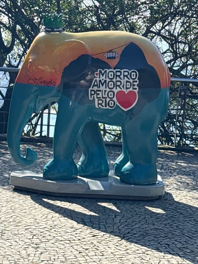 This image features a vibrant, multi-colored elephant sculpture, likely part of an outdoor art installation such as an "Elephant Parade." The sculpture is depicted from a side view, standing on a grey rectangular base on a paved surface. Its body is primarily teal/blue, complemented by sections of orange, tan, and dark blue/black paint.
The most prominent detail on the elephant's side is white text with a red heart symbol. The text reads "MORRO AMOR DE PELO RIO," which translates to "Hill/Favela Love for Rio," strongly indicating a connection to Rio de Janeiro, Brazil, and its characteristic hills or favelas. Below this text, near the trunk, a stylized signature, possibly "Linares," is visible. Towards the rear of the elephant, a small green object resembling a bottle cap displays faint text, including "Mate" and "Refr... Mate," possibly referring to a popular South American beverage.
The setting is outdoors, under bright daylight conditions, as evidenced by the strong, distinct shadows cast on the light-colored cobblestone ground. The shadows suggest a sunny day, likely mid-morning or late afternoon. In the background, the intricate bare branches of trees are visible against a bright sky, indicating an urban park or public space setting. The static nature of the sculpture means no active event is taking place beyond its display as public art.