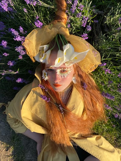 This overhead shot captures a person lying amongst lavender fields in Verkhneye Uchdere, Russia. The subject has long, auburn hair that is partly braided and adorned with white calla lilies, daisies, and lavender sprigs. They are wearing a flowing, bright yellow garment that drapes over them and the surrounding grass and dry soil. The person's face is turned towards the camera, with makeup highlighting their eyes and lips in a way that complements the floral elements. The time of day appears to be late afternoon or early evening, given the warm, directional sunlight casting long shadows and highlighting the textures of the flowers and fabric. The scene evokes a sense of ethereal beauty and tranquility, with the vibrant colors of the flowers and garment contrasting against the natural, earthy setting. There is no visible text in the image.